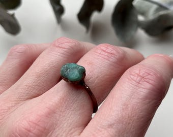 May include: A copper ring with a green gemstone set in the band. The ring is on a person's finger.