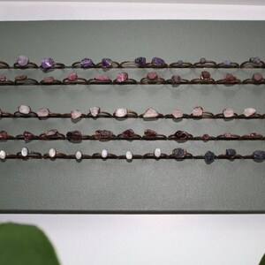 May include: A display of copper wire rings with various semi-precious stones, including amethyst, rose quartz, and green aventurine. The rings are arranged in rows on a green background.