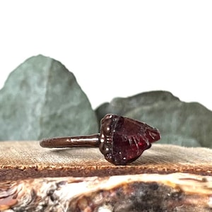 Op de afbeelding: Een ring met een donkerbruine band en een grote, rode, gefacetteerde edelsteen. De edelsteen is gezet in een bezel-setting en heeft een ruwe, natuurlijke uitstraling.