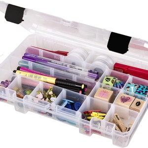 May include: A clear plastic craft organizer with a hinged lid, filled with various craft supplies. The compartments hold pens, washi tape, stamps, buttons, and small embellishments. The organizer is open, showcasing the contents.