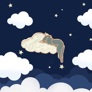 Puede incluir: Un pin de esmalte dorado y azul de un gato durmiendo en una nube blanca con un fondo de noche estrellada.