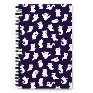 May include: A spiral bound notebook with a purple cover featuring a repeating pattern of white ghost cats. The ghost cats are in various poses, including flying, sitting, and standing.