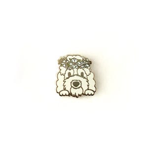 Puede incluir: Pin de esmalte blanco de un perro con una corona de flores. El perro mira al espectador con una expresión feliz.