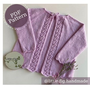 May include: A light purple knitted cardigan with a lace pattern and a tie at the back. The cardigan is laid flat on a white surface with a small wooden sign that says "Little-Fig" and a bunch of dried lavender.