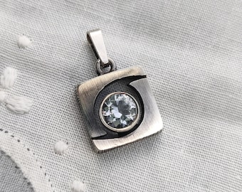 May include: A silver pendant with a square frame and a circular, light blue gemstone. The pendant has a minimalist design and is displayed on a white, textured surface.