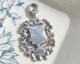 Op de afbeelding: Zilveren bedel met een gedetailleerd, versierd ontwerp in de vorm van een schild. De bedel heeft een lus aan de bovenkant om aan een ketting of armband te bevestigen.