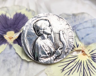 May include: Silver tone metal charm featuring a woman playing a lyre. The charm has the text "Secécille" and "Abrinet" on it.
