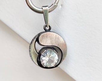 May include: A silver pendant necklace with a round, yin-yang design featuring a clear crystal in the center. The pendant is attached to a silver chain with a lobster clasp.