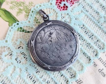 May include: A silver locket with a floral design and a loop for a chain. The locket is on a light blue lace background.