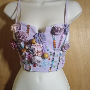 May include: A lavender bustier top with spaghetti straps, adorned with various floral embellishments, beads, and sequins. The decorative elements include fabric flowers, wooden mushroom shapes, and a light blue butterfly. The top is displayed on a white mannequin.