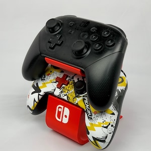 May include: A red Nintendo Switch controller stand with two black controllers. One controller has a Pikachu design and the other is a standard black controller.