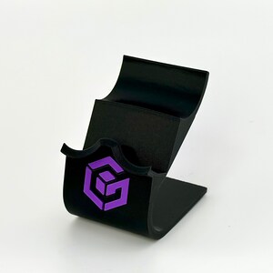 May include: Black phone stand with a purple logo of a cube with the letter G inside. The stand has two slots for phones.