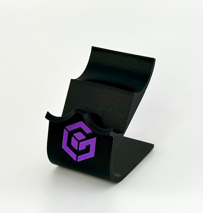 May include: Black phone stand with a purple logo of a cube with the letter G inside. The stand has two slots for phones.