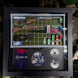 May include: A framed display of Resident Evil 4 video game memorabilia. It includes the game case, two game discs, a shotgun, a rifle, a pistol, a submachine gun, and various other items.
