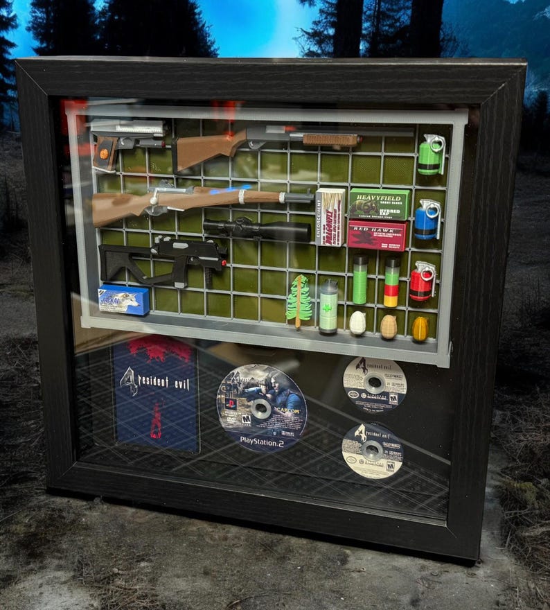 May include: A framed display featuring Resident Evil 4 video game items. The display includes a black and green game case, two game discs, a shotgun, a rifle, a pistol, and various ammunition. The display is set against a green and gray grid background.