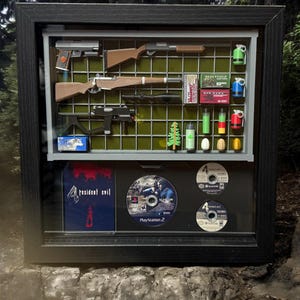 May include: A framed display case with a variety of items from the Resident Evil 4 video game. The case includes a shotgun, a rifle, a pistol, a knife, a grenade, a box of ammo, a game disc, and a game case. The case is made of black wood and has a glass front.