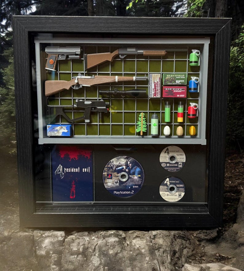 May include: A framed display case with a variety of items from the Resident Evil 4 video game. The case includes a shotgun, a rifle, a pistol, a knife, a grenade, a box of ammo, a game disc, and a game case. The case is made of black wood and has a glass front.