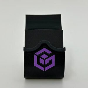 May include: A black phone stand with a purple logo of a cube with the letter G inside. The stand has a curved top and a flat bottom.