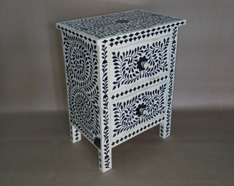 May include: A white nightstand with two drawers and black floral inlay. The nightstand has four legs and is made of wood.