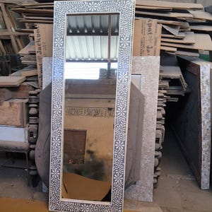 Puede incluir: Un espejo rectangular con un marco blanco con motivos florales. El espejo es alto y se apoya contra una pila de madera y otros objetos. El marco tiene un diseño detallado y el espejo refleja el entorno.