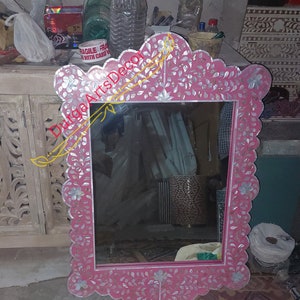 Puede incluir: Un espejo rosa con un marco decorativo hecho de nácar y diseños florales. El marco está tallado de forma intrincada y tiene un borde festoneado.