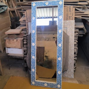 Puede incluir: Un espejo rectangular con marco blanco y un patrón floral azul. El espejo está apoyado contra una pila de madera y otros objetos en un taller. El espejo mide aproximadamente 152 cm de alto.