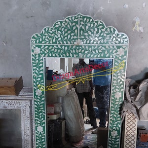 Puede incluir: Un espejo verde y blanco con un diseño floral. El espejo está enmarcado en un marco de madera tallada decorativo. El espejo refleja una habitación con una persona de pie frente a él.