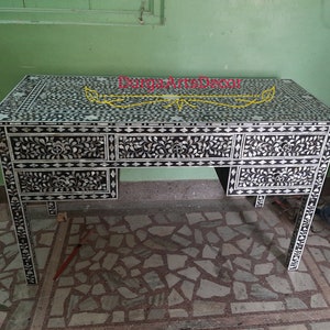 Puede incluir: Un escritorio negro y blanco con intrincados patrones florales incrustados con nácar. El escritorio tiene cuatro cajones y está hecho de madera. El escritorio está sobre un suelo de baldosas.