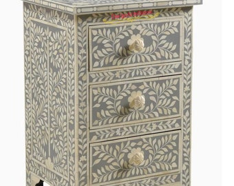 May include: A gray wooden nightstand with three drawers. The drawers have floral bone inlay designs and white bone knobs. The nightstand has a floral bone inlay design on the top and sides.