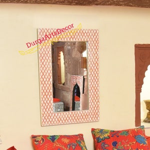 Puede incluir: Un espejo rectangular blanco con un marco de patrón geométrico rojo y blanco. El espejo está colgado en una pared con un fondo crema. El espejo refleja una habitación con un arco similar y un cojín de patrón rojo y blanco.