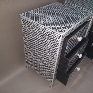 May include: A black and white bone inlay nightstand with three drawers. The drawers have black fronts and bone inlay handles. The nightstand has a geometric pattern.