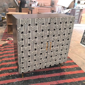 Puede incluir: Un mueble con un patrón geométrico en blanco y negro, tiradores y patas doradas. El mueble de madera está sobre una alfombra a rayas rojas y negras. El mueble mide unos 100 cm de alto.