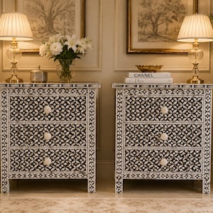 Bone inlay Two white bedside tables with three drawers each. The drawers feature black and white geometric patterns and white knobs. The bedside tables are made of wood and have a distressed finish.