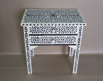May include: A white end table with two drawers and black floral inlay. The table has four legs with black floral inlay.