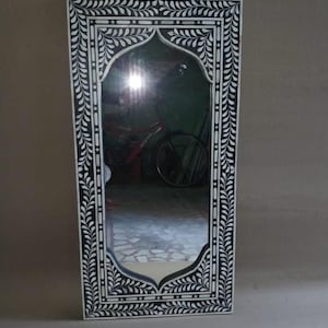 Puede incluir: Un espejo de incrustaciones de hueso negro y blanco con un diseño floral decorativo. El espejo tiene una parte superior arqueada y está enmarcado en un marco rectangular.