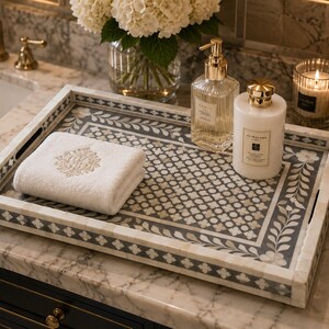 Luxury marble bathroom vanity featuring an ornate bone inlay tray with floral motifs, designer toiletries, folded white towel, candlelight, white hydrangeas, and gold fixtures in a high-end spa-inspired setting.