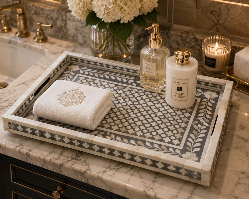 Luxury marble bathroom vanity featuring an ornate bone inlay tray with floral motifs, designer toiletries, folded white towel, candlelight, white hydrangeas, and gold fixtures in a high-end spa-inspired setting.