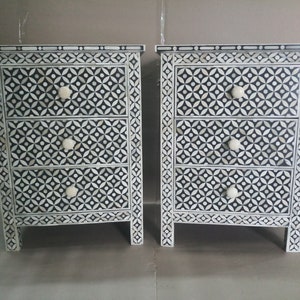 May include: Two black and white nightstands with three drawers each. The drawers have a geometric pattern and white knobs. The nightstands are made of wood and have a traditional style.