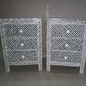 May include: Bone inlay Two white and black geometric patterned bedside tables with three drawers each. The drawers have white knobs.