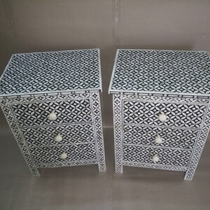 May include: Bone inlay Two white bedside tables with three drawers each. The drawers feature black and white geometric patterns and white knobs. The bedside tables are made of wood and have a distressed finish.