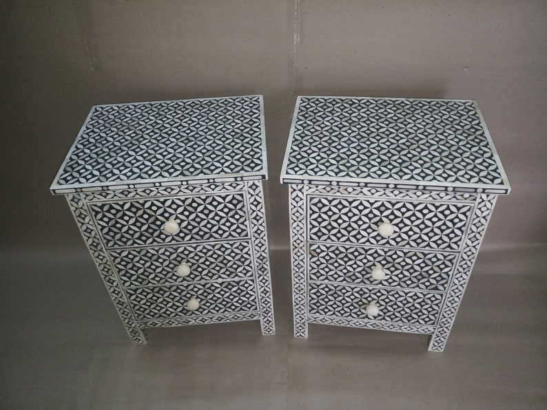 May include: Bone inlay Two white bedside tables with three drawers each. The drawers feature black and white geometric patterns and white knobs. The bedside tables are made of wood and have a distressed finish.