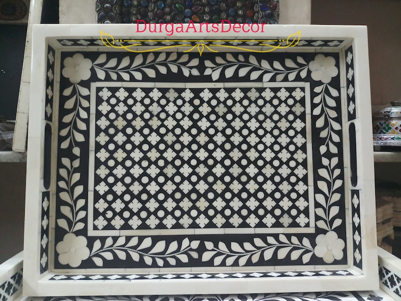 K&ouml;nnte beinhalten: Ein rechteckiges Serviertablett mit einem geometrischen Muster in Schwarz und Wei&szlig; aus Knocheninlay. Das Tablett hat einen dekorativen Rand mit floralen Motiven.