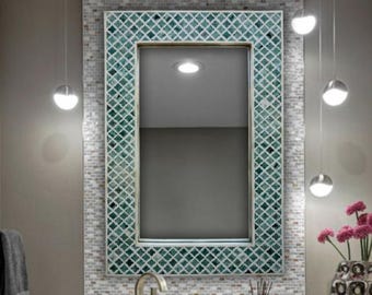 May include: A bathroom mirror with a decorative green and white mosaic tile frame. The mirror is mounted on a mosaic tile wall, with a brushed nickel faucet and sink. Two pendant lights hang above the mirror, and a vase of pink flowers sits on the counter.
