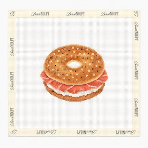 Può includere: Una tela per ricamo con un bagel realizzato a punto croce con formaggio spalmabile e salmone. Il disegno è centrato su una griglia bianca con il testo BirchNDLPT stampato ripetutamente lungo il bordo color crema della tela quadrata.