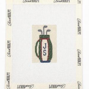 May include: A needlepoint canvas featuring a centered design of a green golf bag with red trim and blue accents, containing three golf clubs. The letters JCS are stitched in blue on the bag. The border is printed with the text Birch NDLPT.
