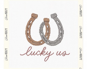May include: A cross-stitch pattern on white fabric featuring two overlapping horseshoes, one in brown and one in gray, with the text "lucky us" stitched in red cursive below. The border of the fabric is printed with the text "Birch NDLPT".