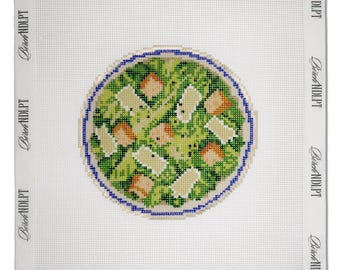 May include: A square needlepoint canvas featuring a circular design of a green salad with croutons in a blue-rimmed bowl. The white canvas border is printed with the repeating text Birch NDLPT along all four edges of the fabric.