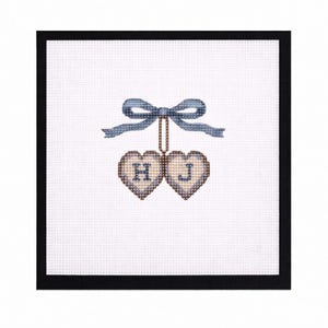 Può includere: Un disegno a punto croce con due ornamenti a forma di cuore con le lettere "H" e "J" ricamate. I cuori sono sospesi a un fiocco blu. Il disegno è ambientato su uno sfondo bianco con un bordo nero.