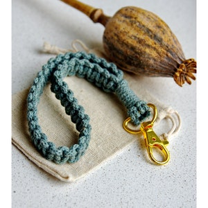 May include: A teal blue braided keychain with a gold metal clasp. The keychain is lying on a small, off-white fabric pouch.