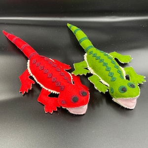 Peut inclure: Deux lézards en peluche en feutre, l'un rouge et l'autre vert. Le lézard rouge a une bordure blanche et des yeux noirs. Le lézard vert a une bordure blanche et des yeux verts.